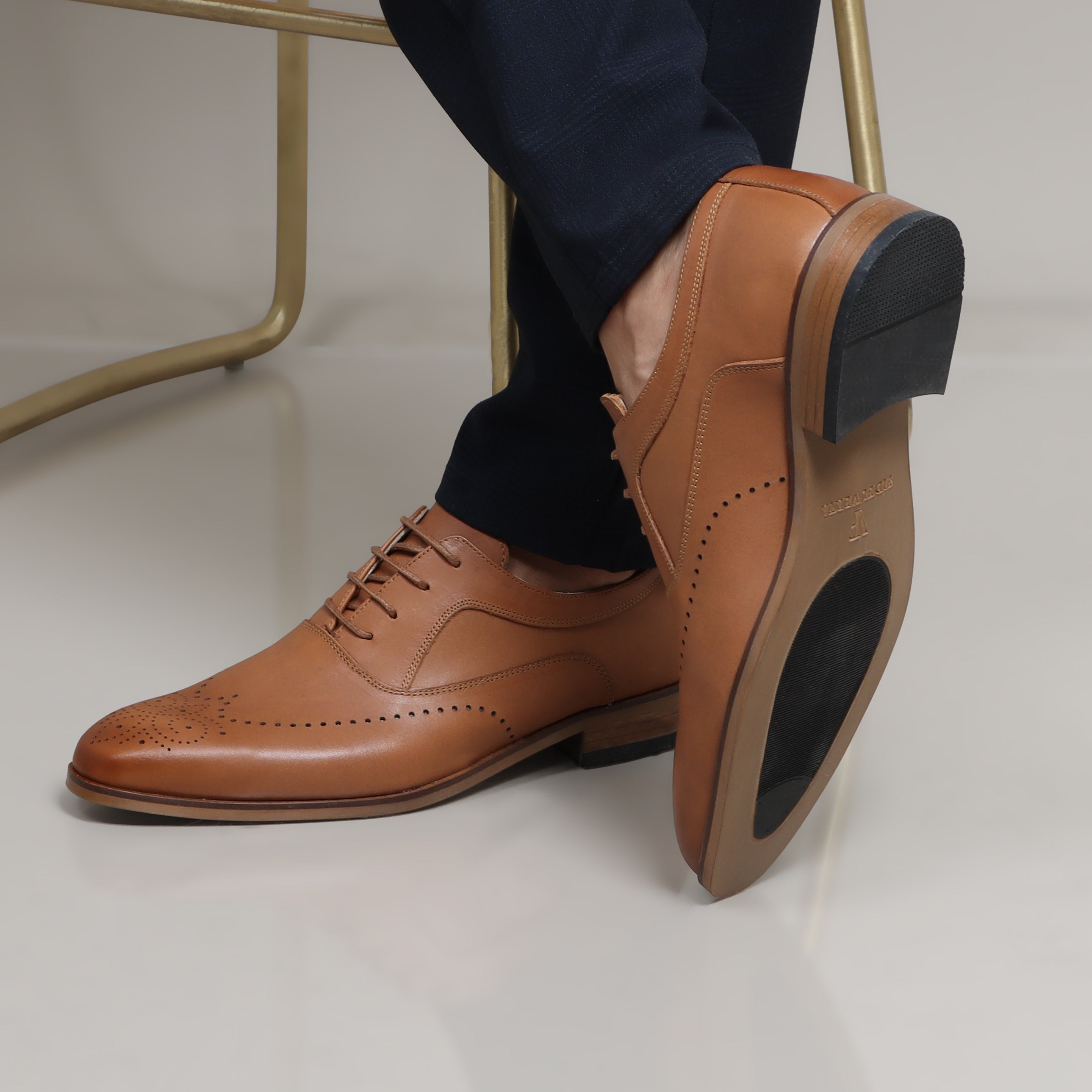 Tan leather Oxford shoes with brogue details and black rubber soles worn with navy trousers.