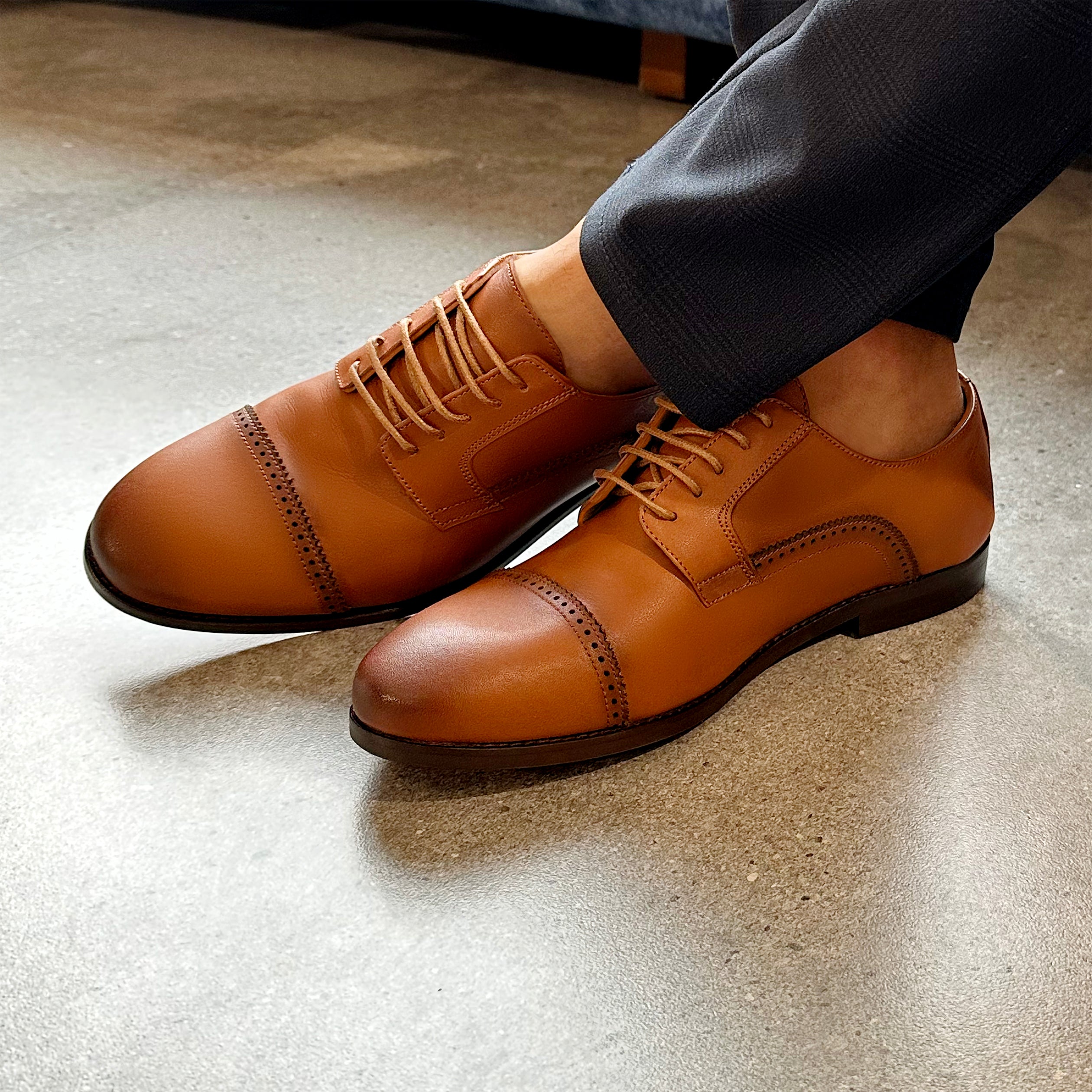 Brown leather Oxford shoes with cap toe stitching and slim laces on model wearing black pants.
