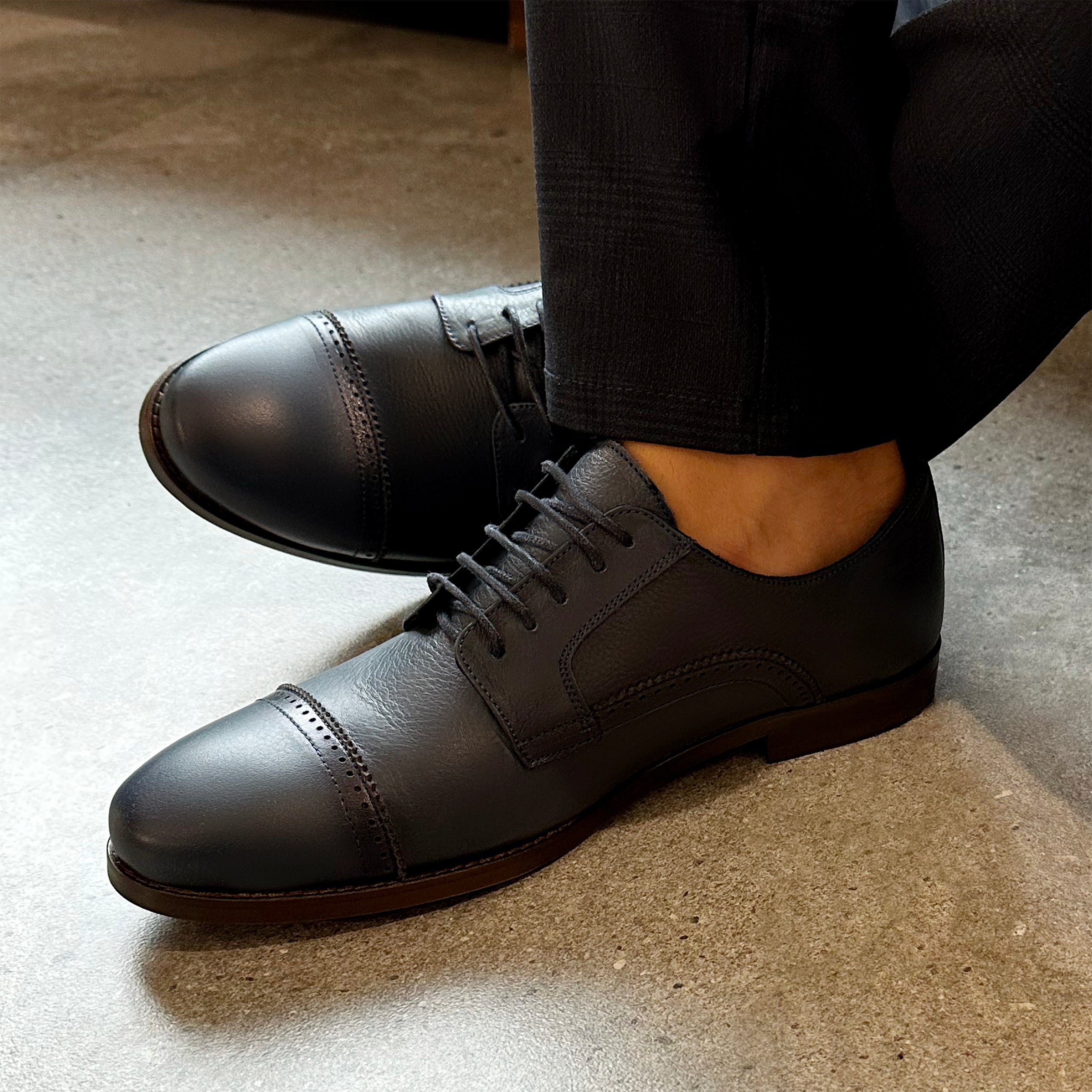 Navy leather Oxford shoes with cap toe and closed lacing worn with black trousers.