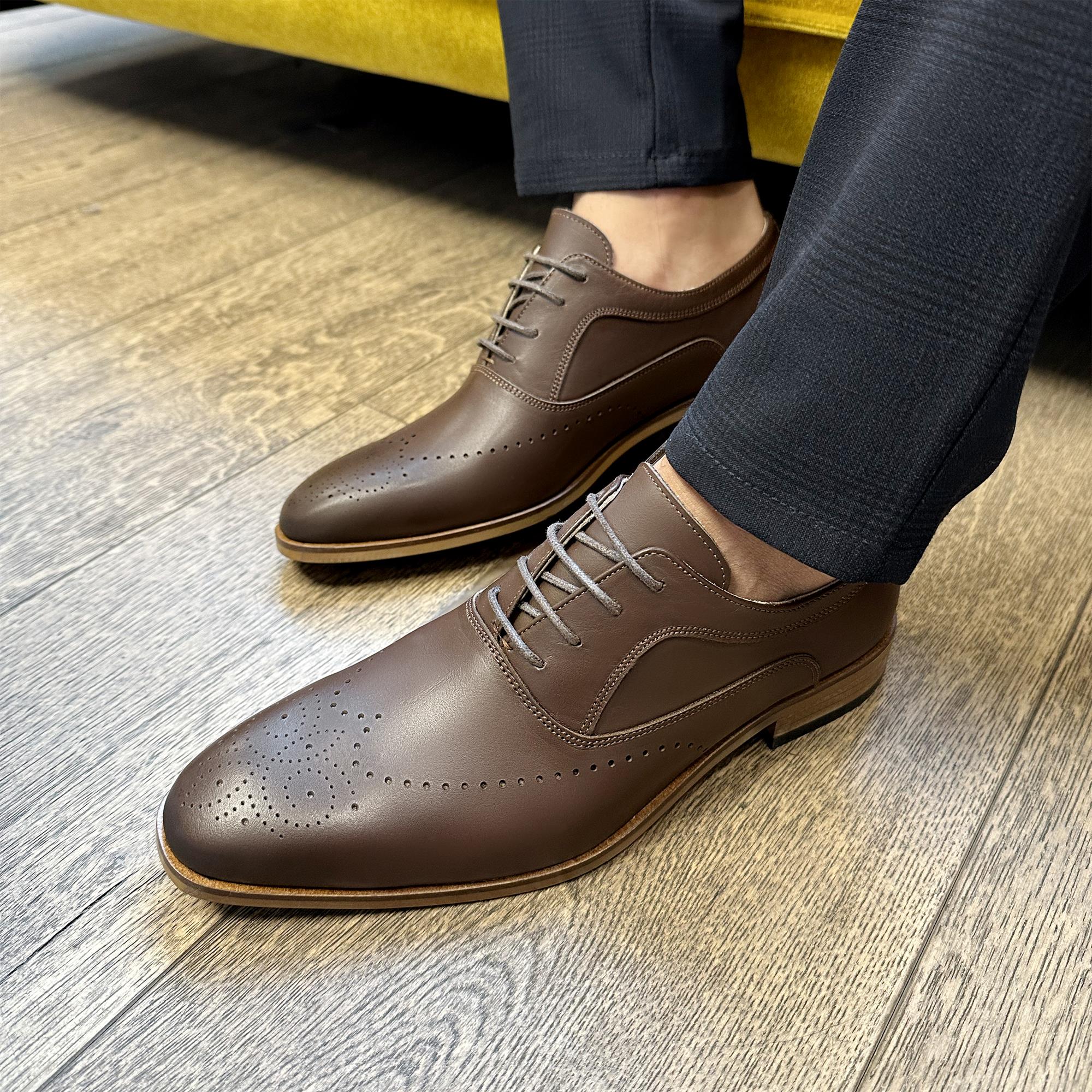 Brown leather English classic shoes with brogue detailing and lace-up closure worn with black trousers.