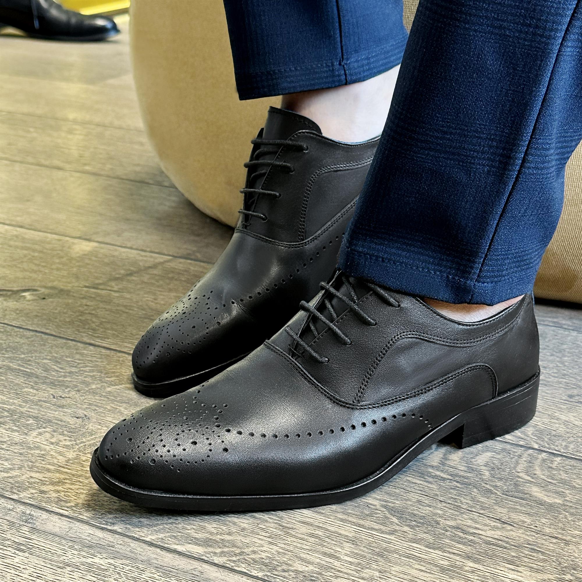 Black leather classic English shoes with brogue detailing and laces worn with navy blue trousers.